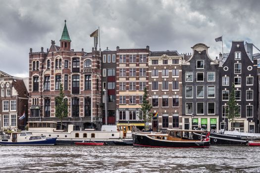 Amsterdam, the Netherlands - July 1, 2019: Line of historic houses with gables along Amstel River downtown. Many houseboats and other vessels on dark river under heavy cloudscape.
