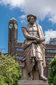 Amsterdam, the Netherlands - July 1, 2019: Beige full body of Rembrandt van Rijn statue on Rembrandtplein under blue sky with white clouds. Clock tower in brown stones in back.