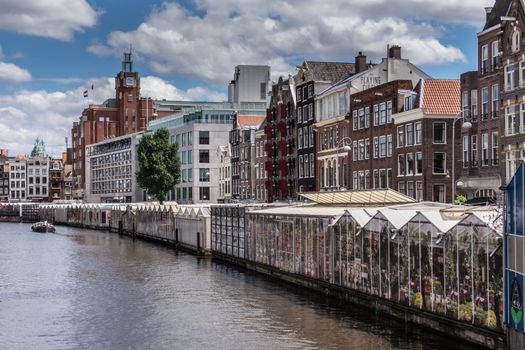 Amsterdam, the Netherlands - July 1, 2019: Long row of glasshouses built on barges moored on Singel Canal and housing flower market. Back is row of houses under blue cloudscape,