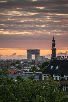 Amsterdam, the Netherlands - July 2, 2019: Yellow and red lights cut by dark clouds in sky over towers and roofs. Westerkerk spire. Modern Pontsteiger high rise.