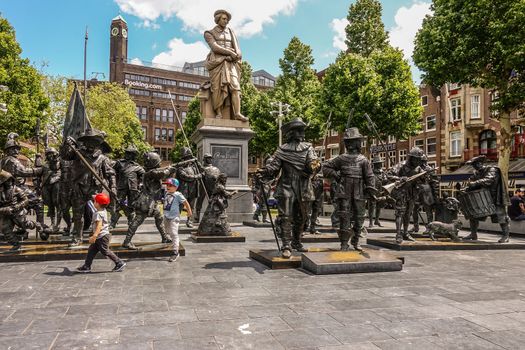 Amsterdam, the Netherlands - July 1, 2019: De Nachtwacht compostion of statues on Rembrandtplein in front of beige tall statue of Rembrandt Van Rijn, with booking.com office buildign in back.