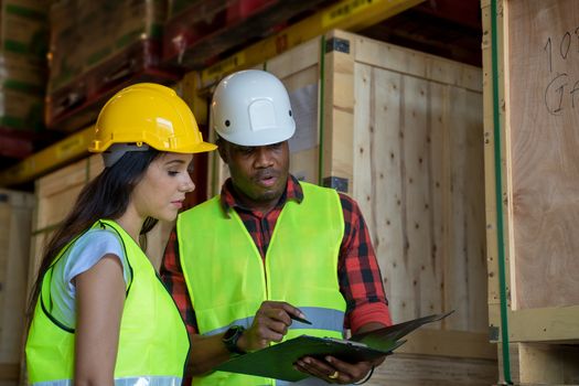 Warehouse manager and warehouse worker wearing helmet  with clipboard checking products about delivery schedule in industrial storage room.