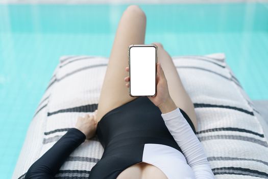 Woman in black and white swimsuit holding mockup blank mobile phone with white screen near pool.