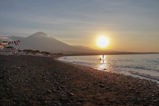 People enjoying surf and paddleboarding at sunset on a tropical island. Panorama with volcano and mountain range. Fishing boats parked on the beach.