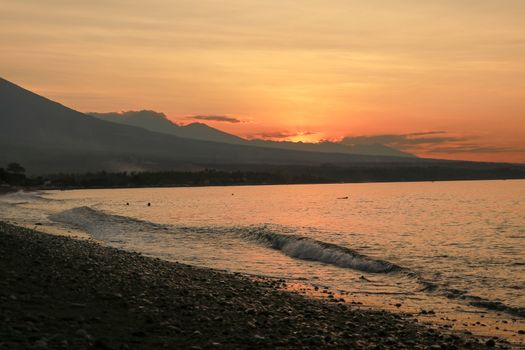 Orange sunset on tropical island. Calm sea lit by the rays of the setting sun. Panorama with volcano and mountain range. Fishing boats anchor on beach.