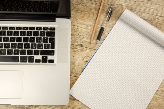 Contrast between old and new office work: shot from the top of an aluminum laptop on a wooden desk with an open checkered notebook, a pencil and a pen