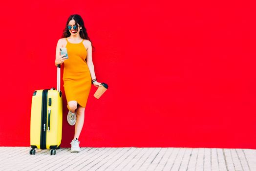 traveling woman with a yellow suitcase using a smartphone and drinking a coffee.