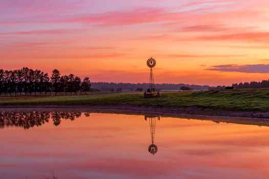 Windmill sits on farm land near a dam with beautiful sunrise sky and water reflections.  A bird sits on one of the fins of the windmill