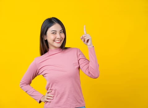Asian smiling young woman finger Pointing up for advertise, banner or showing on isolated yellow color background, wearing winter cozy sweater indoors studio, happiness and fun, copy space concept