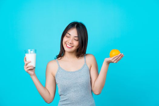 Attractive Asian young woman holding a glass of milk and a orange on isolated blue color background, weight loss and avoid junk food for dieting and Healthy