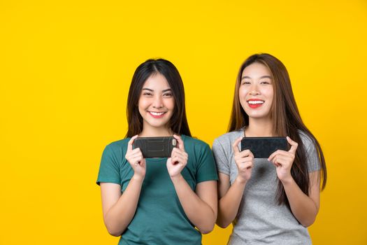 Two Happiness Asian smiling young woman gamer using smart mobile phone and playing games on isolated yellow color background, Lifestyle and leisure with hobby concept