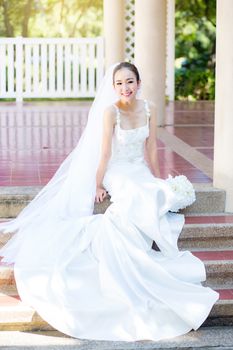 bride is sitting with flowers. Beautiful Young woman posing in park or garden in white bridal dress outdoors on a bright sunny day.