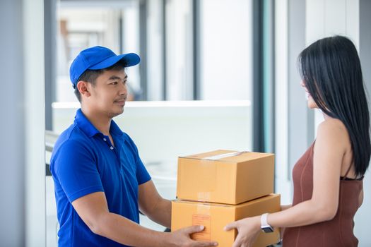 Asian delivery servicemen wearing a blue uniform with a bluecap and  handling cardboard boxes to give to the female customer in front of the house. Online shopping and Express delivery