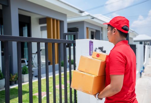 Asian delivery servicemen wearing a red uniform with a red cap and handling cardboard boxes to give to the female customer in front of the house. Online shopping and Express delivery