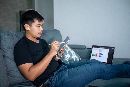 Asian businessman using wireless earphones and using computer notebook and tablet working from home.He is prepare graphs and information for online meetings at home.