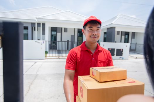 Asian delivery servicemen wearing a red uniform with a red cap and handling cardboard boxes to give to the female customer in front of the house. Online shopping and Express delivery