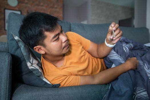 Asian man is ill and coughs on the sofa at home and he uses a thermometer to check the temperature during the coronavirus epidemic.