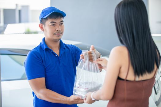 Asian delivery servicemen wearing a blue uniform with a blue cap and handling food boxes in plastic bags to give to the customer in front of the house. Online shopping and Express delivery