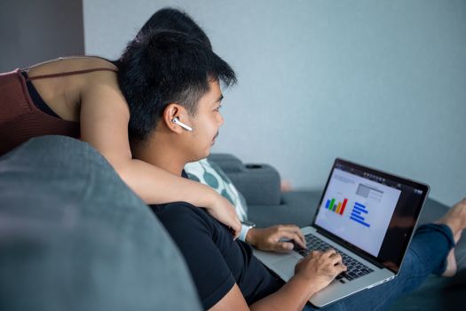 Asian young smiling couple hugging and using wireless earphones and using computer notebook working from home .He is prepare graphs and information for online meetings at home.