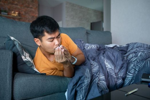 Asian man is ill and coughs on the sofa at home and he uses a thermometer to check the temperature during the coronavirus epidemic.