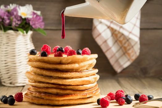 Syrup pouring a stack of pancakes with berries on a wooden table in a rustic style.