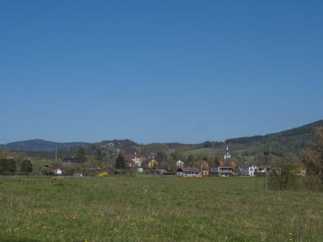 Spring landscape with view on village Cvikov in Lusitian mountains with old and modern houses and lush green grass meadow, deciduous and spruce tree forest and hills, blue sky background.