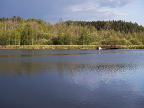 view on calm water of forest lake, fish pond Kunraticky rybnik with dam pier, birch and spruce trees growing along the shore and clear blue sky in golden sun light. Nature background. Spring landscape.