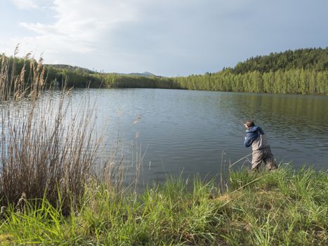 Fly Fisherman angler standing on shore of calm water of forest lake, fish pond Kunraticky rybnik with birch and spruce trees growing along the shore. Nature fishing background. Springtime.