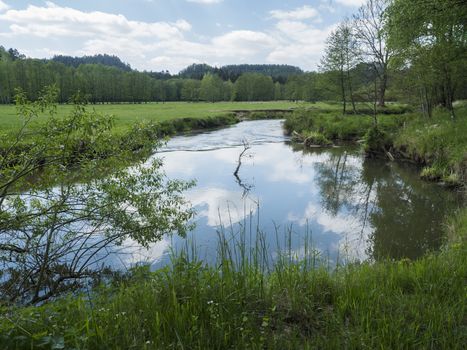 idyllic landscape of winding river stream meander at lush green meadow with deciduous tree forest, blue sky backgound. Late spring sunny afternoon, vibrant colors.