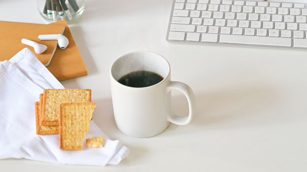 Top view of coffee cup, biscuits, white button keyboard, pencil holder, notebook and white napkin on white desk.