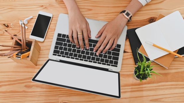 Top view of woman hand while using/typing on black blank screen laptop including pencil holder, black blank screen smartphone, potted plant, pencil, notes, earphone on wooden desk.
