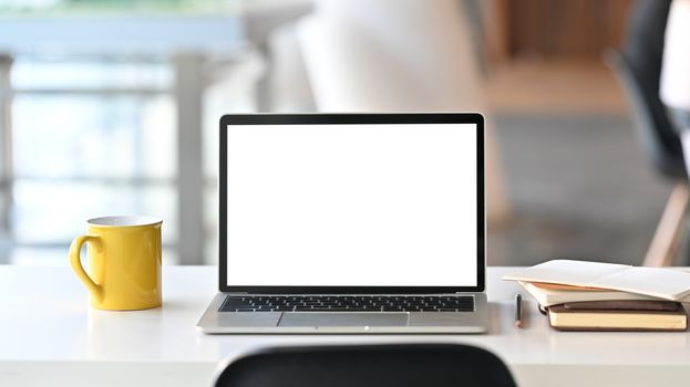 Laptop with white blank screen display and office equipment are putting on the white office desk over the modern office background.