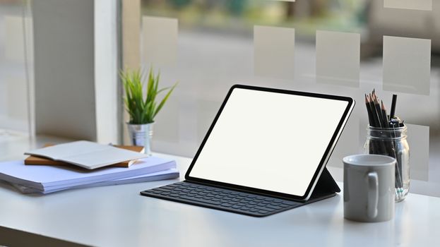 Photo of white blank screen computer tablet in a leather-case and keyboard is setting on a modern working table over comfortable office background.