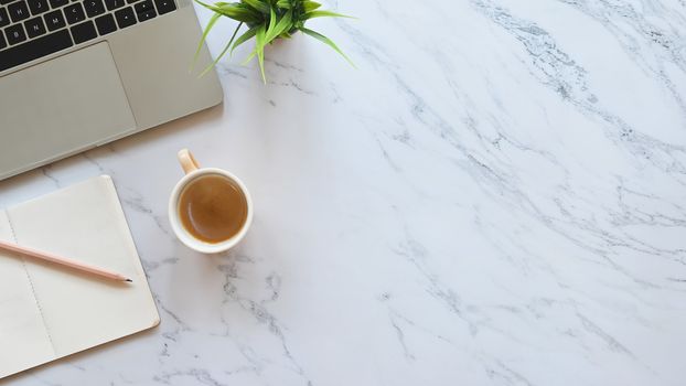 Close-up image part of laptop including coffee cup, pencil, notebook and potted plant putting on marble office table.