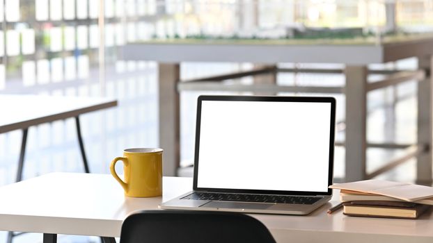 Modern laptop with white blank screen display and office equipment are putting on the white office desk over the modern office background.