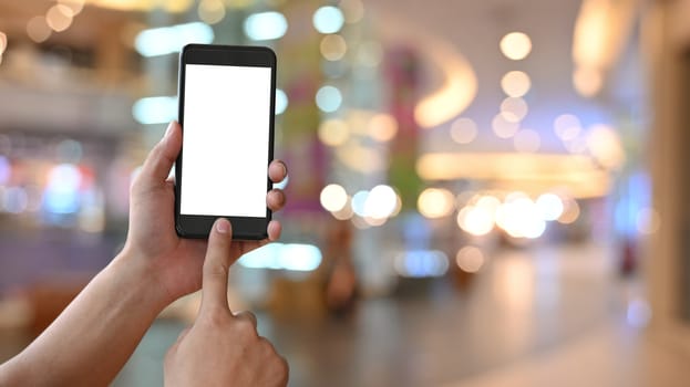 Close-up image of hand while holding crop black smartphone with white blank screen display and touching on home button over the shopping center blurred background.