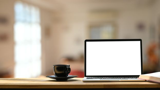 Photo of white blank screen laptop, black coffee cup and coasters on the wooden working desk over blurred modern cafe background.