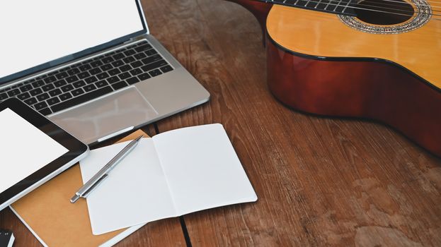Work table of songwriter. Cropped shot of note, pencil, acoustic guitar, white blank screen tablet and laptop all of these is putting on the wooden desk.