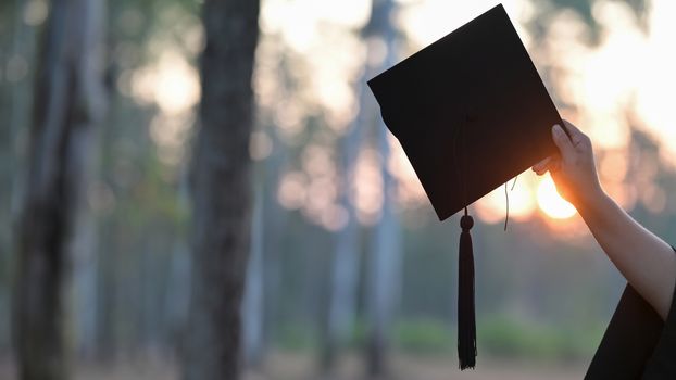 Graduate woman hand holding the graduation cap in her hand over blurred forest background.  Graduation celebration concept.