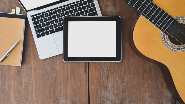 Working desk of songwriter. Top view of note, pencil, laptop, white blank screen tablet and acoustic guitar all of these is putting on the wooden desk.
