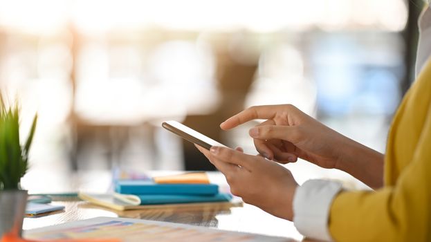 Side view of cheerful designer using black smartphone on hand and pointing finger on it while sitting at the wooden working desk.