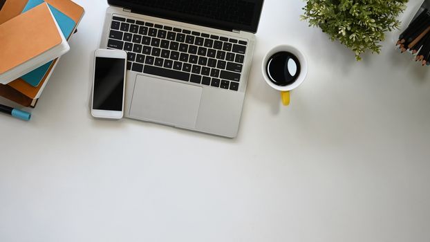 Modern office working desk including a coffee cup, laptop, black blank screen smartphone, potted plant, pencil and books putting on it. Office equipment with flat laid.
