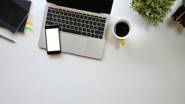 Top view office desk including a coffee cup, laptop, white blank screen smartphone, potted plant, pen and notebook putting on it. Office equipment with flat laid.
