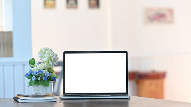 White blank screen laptop, notebook, smartphone and potted plant all of these putting together on the modern working desk with comfortable living room as background.