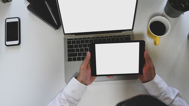 Top view image of business man holding a white blank screen tablet at the modern working desk with the white blank screen laptop and coffee cup, smartphone, notebook and potted plant as background.