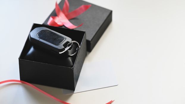 Close-up image of the Digital car key putting inside the black gift box with red ribbon and wish card on the white desk as background. Surprising Valentine's Day gift concept.