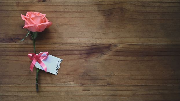 Top view image of pink rose and wishes card tied together with red ribbon all of these putting on the wooden table. Surprising Valentine's Day gift concept.