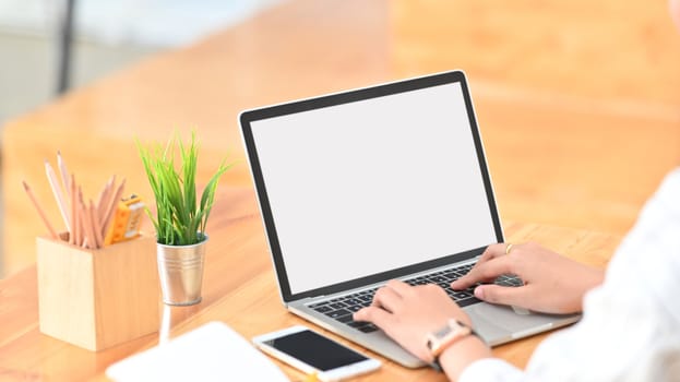 Cropped shot of young creative woman surfing the internet by using white blank screen laptop while sitting at the modern wooden desk.