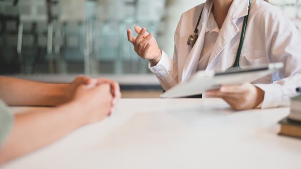 Cropped shot of woman working doctor while giving an illness information to her patient at the white working desk with the modern hospital as background.