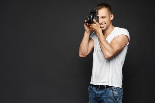 Young smiling handsome man holding a vintage photo camera and making a photoshoot, isolated at the dark background.